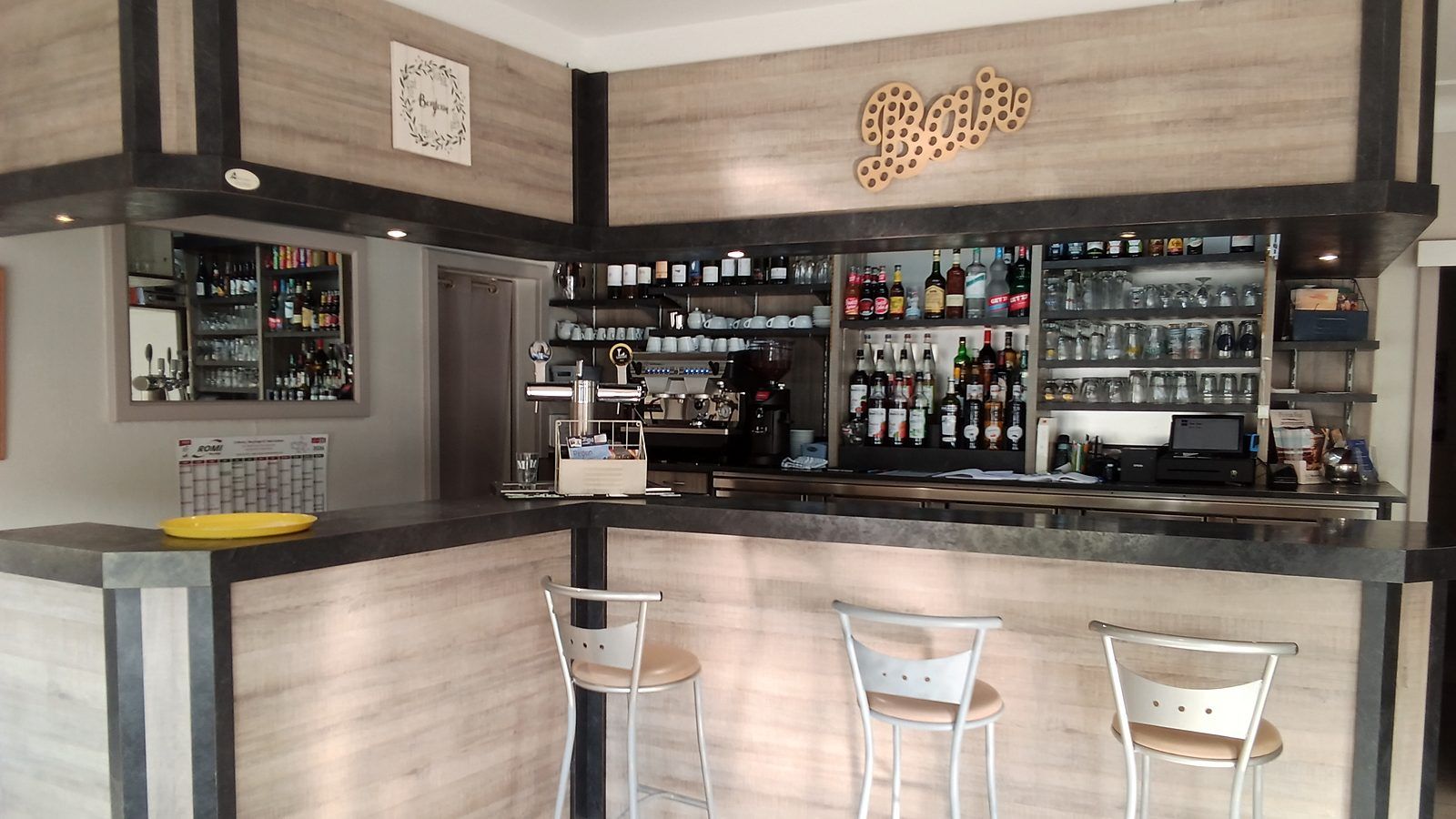 Le comptoir du bar en angle, ses trois tabourets hauts, la machine à café et la tireuse ; derrière, les étagères de bouteilles et de verres sous l’enseigne « Bar » en lettres de bois.