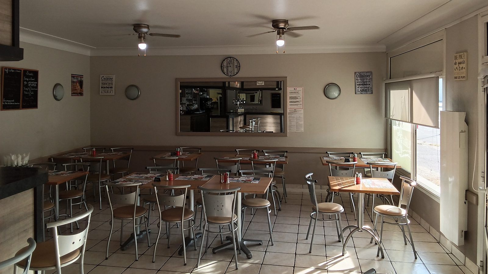 La salle de restaurant et ses tables carrées dressées de sets de table, sous deux ventilateurs de plafond ; au fond, l’ardoise des desserts et le passe de la cuisine, et à droite la fenêtre sur la rue.