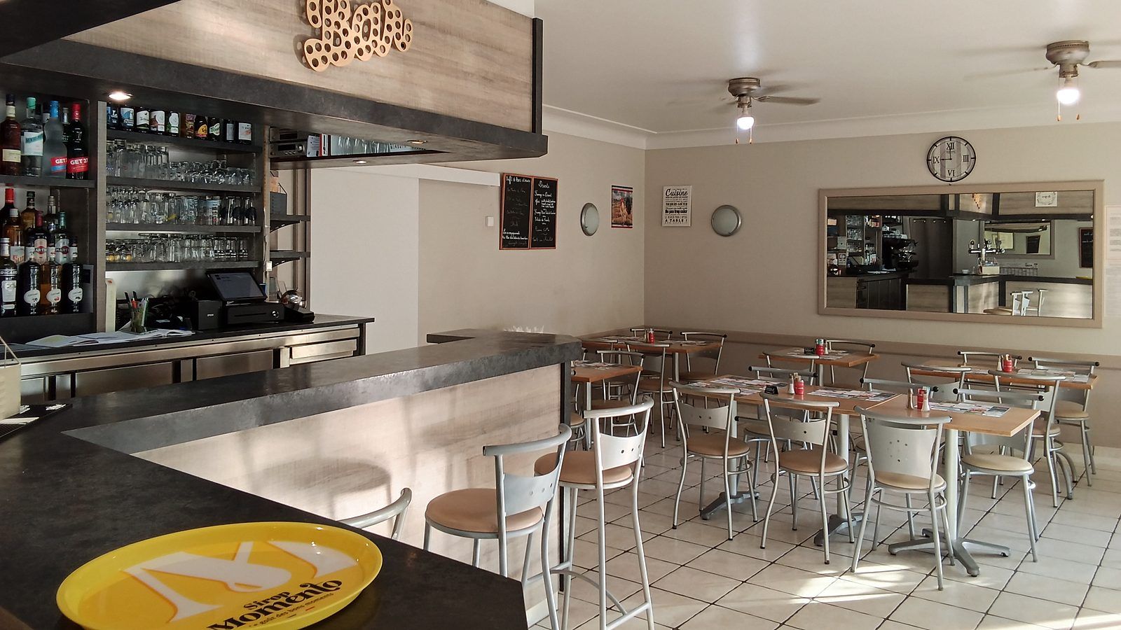 La salle vue depuis le comptoir : le bar et ses bouteilles au premier plan à gauche, les tables et leurs chaises qui s’étendent vers le fond, et l’horloge murale au-dessus.